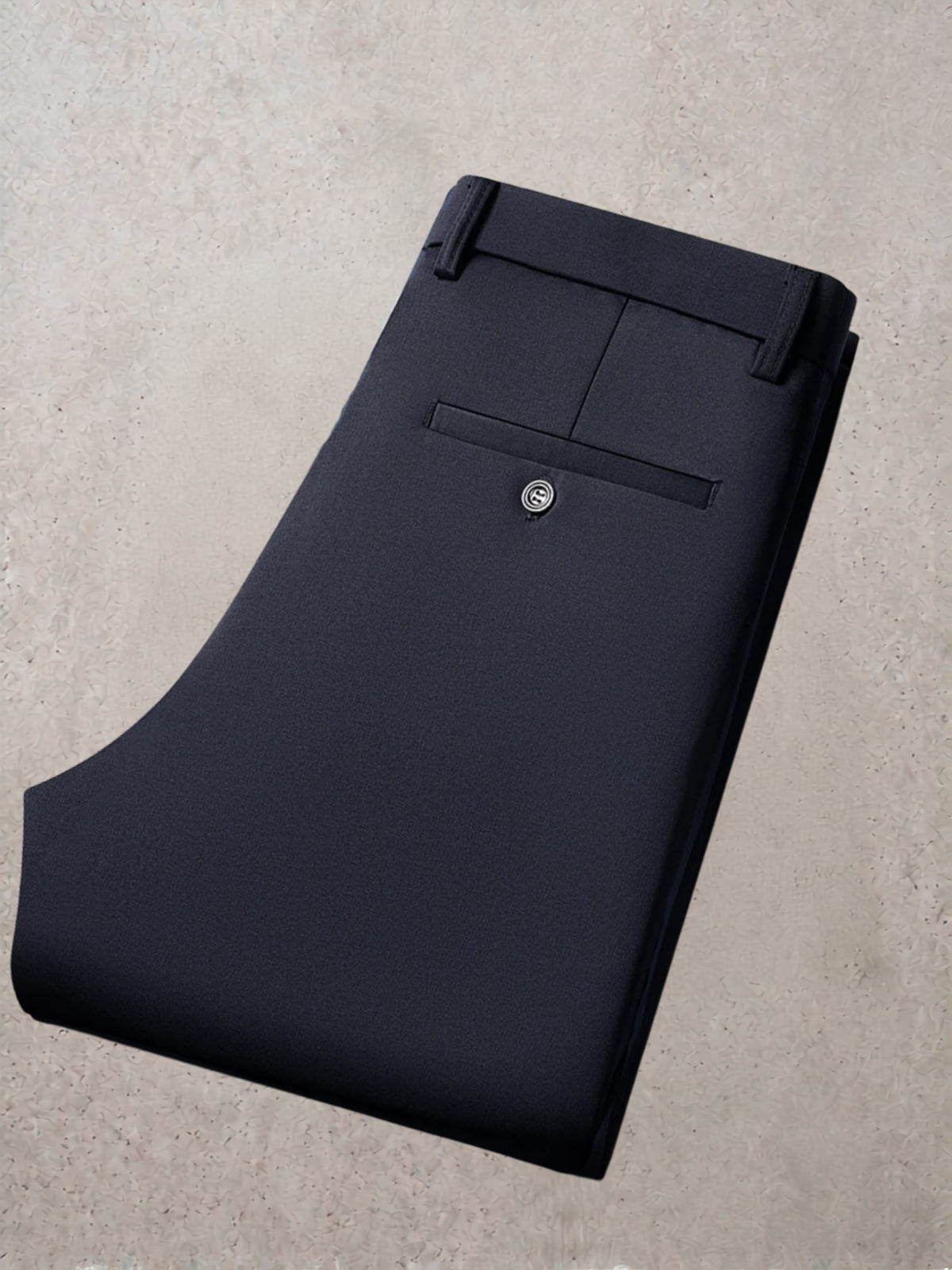 A folded pair of dark navy trousers displayed on a textured light beige concrete surface, showing the waistband, belt loops, and a single button closure on the back pocket.